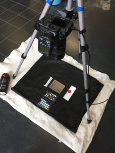 Image of a digital camera on a tripod pointed down to the ground where a roman tablet, color pallet, and small red ball have been placed.